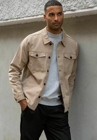 Vīrietis piespiedies pie pelēkas sienas, valkājot bēša krāsas pogājam džemperi virs pelēka sporta krekla un melnas bikses, ar rokām kabatās.