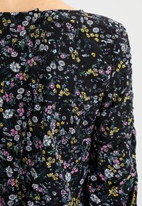 Svart texturerad tygblus med ett flerfärgat blommönster i vitt, rosa och gult, som fästs med en liten knapp baktill.