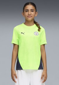 Ragazza con lunghi capelli intrecciati che indossa una maglietta da calcio neon verde del Manchester City e pantaloncini bianchi, in piedi su uno sfondo grigio.