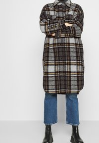 Persona che indossa un lungo cappotto a scacchi sopra jeans blu e stivali neri, in piedi con le braccia incrociate contro uno sfondo chiaro e semplice.