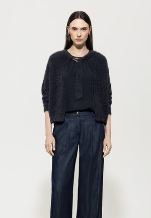 Marineblaue, flauschige Strickjacke mit rundem Ausschnitt und geknotetem Verschluss, schichtweise über einem dunklen Oberteil getragen und kombiniert mit lockeren, weit geschnittenen Jeans mit dunkler Waschung.