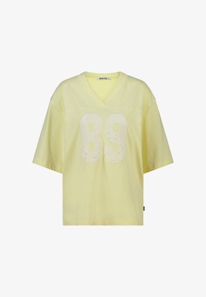 Chemise ample à manches courtes avec col en V, de couleur jaune pâle, arborant un grand chiffre texturé "89" sur le devant et une petite étiquette sur le côté.
