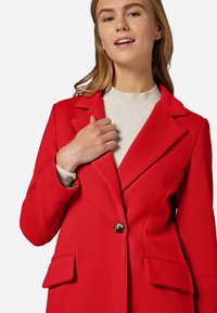 Blazer rouge en tissu structuré avec des revers notched, fermeture à un bouton, poches avant doubles et motif texturé subtil.