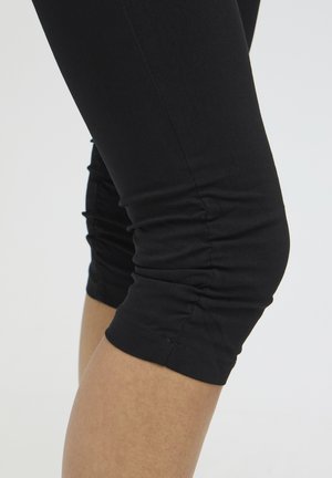 Detailný záber na ohnutú nohu obutú v čiernych zvrásnených skrátených legínach nad lýtkom na jednoduchom svetlom pozadí.