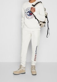 Felpa bianca in cotone con logo Timberland, abbinata a pantaloni della tuta coordinati con dettagli del logo. Stivali da trekking marroni, zaino beige con spallacci neri.