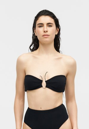 Νεαρή γυναίκα με βρεγμένα σκούρα μαλλιά, φορώντας μαύρο μπικίνι χωρίς τιράντες με κεντρικό μεταλλικό κρίκο και εσώρουχα με ψηλή μέση, μπροστά σε απλό φόντο.