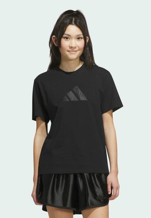 Ung kvinde iført sort Adidas t-shirt og skinnende sorte shorts, stående mod en ensfarvet lys baggrund med fronten mod kameraet.