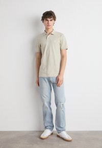 Ανοιχτό μπεζ πόλο μπλουζάκι με απαλή υφή, κοντά μανίκια και διακριτό λογότυπο; συνδυασμένο με ανοιχτό μπλε τζιν με φθορές και λευκά αθλητικά παπούτσια.