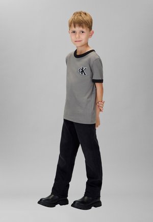 Grijs T-shirt met korte mouwen met zwarte accenten en een 'CK' patch, gecombineerd met zwarte broek en zwarte enkellaarzen.