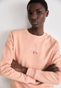 Sudadera de color durazno claro con cuello de tripa y puños acanalados. Presenta un logo bordado en blanco de Calvin Klein en el pecho. Textura de tela suave.