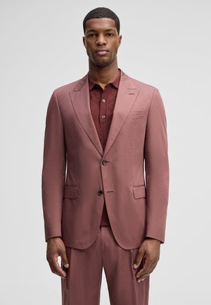 Mężczyzna w różowym garniturze w odcieniu dusty rose i pasującej bordowej koszulce polo z wzorem, stojący na jasnoszarym, jednolitym tle.