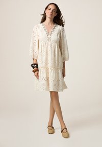 Vestido blanco bordado con escote en V, largas mangas globo y dobladillo en capas. Realzado con un intrincado bordado alrededor del escote.