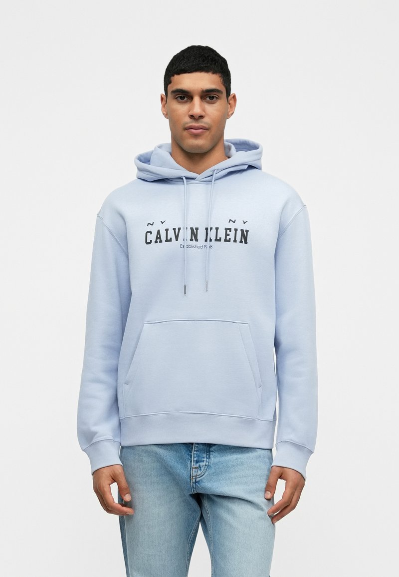 Svetlomodrá mikina s kapucňou a predným vreckom, sťahovací šnúrkový golier a čierny potlač "Calvin Klein". Jemný bavlnený zmesový materiál, voľný strih.