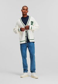 Cárdigán crema con acentos en verde y negro, gran aplicación de número en la parte delantera, combinado con camiseta azul oscuro y jeans azul claros. Tela texturizada.