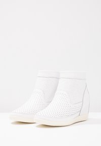 Bottes blanches à la cheville avec une texture perforée, dotées d'un talon compensé et d'accents lisses. Le design est moderne, mettant l'accent sur le minimalisme.