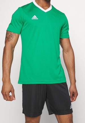 Mężczyzna w zielonej sportowej koszulce Adidas z białym kołnierzykiem i czarnych szortach Adidas, stojący z rękami swobodnie opuszczonymi wzdłuż ciała.