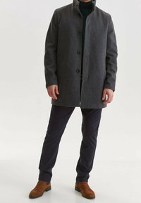 Grauer Wollmantel mit hohem Kragen, Knopfverschluss und Seitentaschen, kombiniert mit dunkler Hose und braunen Wildleder-Stiefeletten.