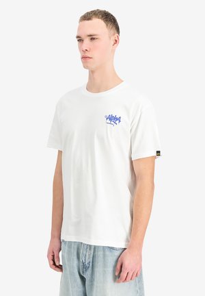 Junger Mann in weißem T-Shirt mit blauem "Alpha Industries"-Logo auf der Brust, trägt hellblaue Jeans, steht vor weißem Hintergrund.