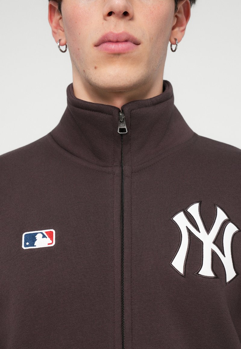 Mørkebrun zip-up jakke med et hvidt New York Yankees-logo og et lille MLB-patch, med en glat tekstur og høj krave.