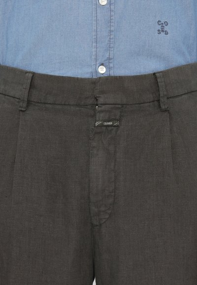 Svart linnebyxor med platt framdel, pryds av en liten etikett med ordet "CLOSED." Matchas med en blå chambray knäppskjorta.