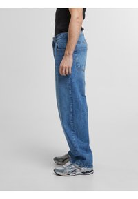 Jeans en denim bleu clair avec une coupe décontractée, un design à cinq poches et des détails de couture visibles. Associés à des chaussures de sport grises et bleues.