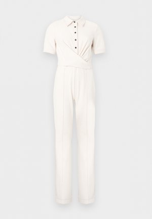 Ženska z dolgimi temnimi lasmi, oblečena v kratkoročni kombinezon v slonokoščeni barvi s pasom, ki se veže ob strani, in nosi bež sandale z odprtimi prsti, stoji pred enobarvnim ozadjem.