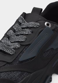 Gros plan sur une chaussure de sport noire et grise montrant des lacets texturés, une tige en tissu mélangé et synthétique, ainsi que des œillets en métal.