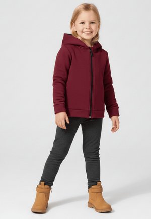 Uśmiechnięte dziecko stojące, ubrane w bordową bluzę na zamek, ciemnoszare legginsy i beżowe botki na kostkę na jednolitym jasnym tle.