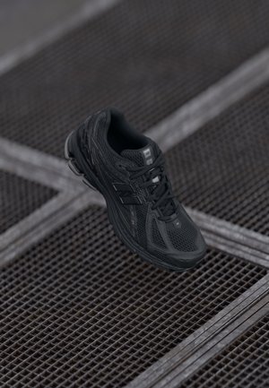 Black athletic shoe with mesh upper, textured surface, lace-up design, and cushioned sole. Positioned above a metal grid background.