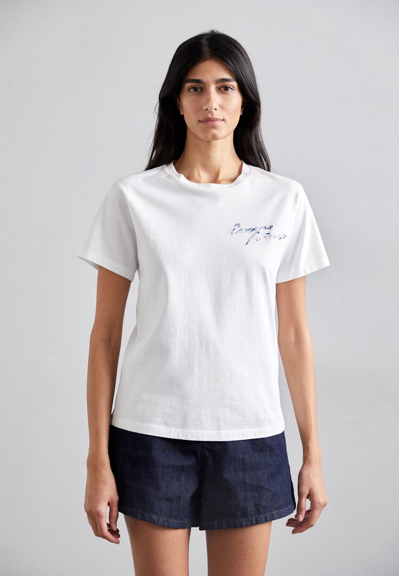 Weißes Baumwoll-T-Shirt mit Rundhalsausschnitt, das auf der linken Seite blau gestickten Text aufweist. Kombiniert mit dunklen Denim-Shorts.