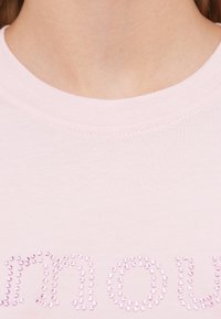 Ροζ ανοιχτό t-shirt με στρογγυλή λαιμόκοψη και διακοσμητικό κείμενο από στρας. Το ύφασμα φαίνεται απαλό με εφαρμοστό σχέδιο.