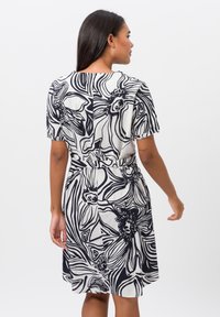 Korte mouwen jurk in het wit met zwarte bloemenpatronen. Beschikt over een ceintuur op de taille en een vloeiende silhouet. Zachte stof textuur.
