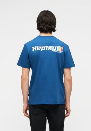 Homme aux cheveux foncés portant un T-shirt bleu avec le texte « Replay Racing Team 1981 » et un logo rayé sur le haut du dos, associé à un pantalon noir.