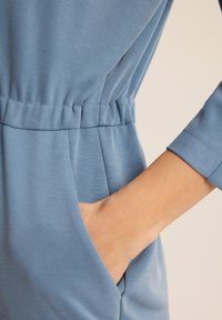 Jasnoniebieski ciuch z wciągniętą talią, wygodny materiał i boczna kieszeń. Mankiety rękawów są proste, co dopełnia cały projekt.