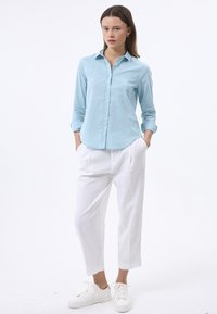 Camisa de manga larga azul claro con cuello abotonado, combinada con pantalones de lino blancos holgados y zapatillas blancas. La camisa tiene una tela texturizada.