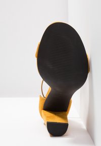 Żółte zamszowe sandały na wysokim obcasie, z kwadratowym noskiem i paskiem wokół kostki. Podeszwa jest czarna i ma teksturowany, prążkowany wzór.