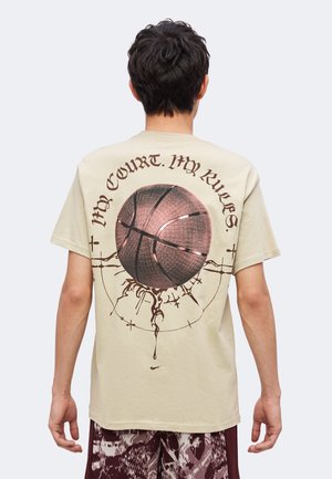 Mann, der ein beigefarbenes T-Shirt mit einer großen Grafik eines Basketballs und dem Text „My Court. My Rules“ auf dem Rücken trägt.