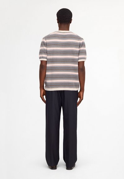Mann steht mit dem Rücken zur Kamera, trägt ein kurzärmeliges, gestreiftes Strickshirt und lockere schwarze Nadelstreifenhose vor weißem Hintergrund.