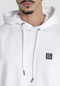 Biela mikina z jemného materiálu, s prednou kapsou, šnúrkami s kovovými akcentmi a malou čiernou nášivkou.