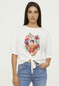 T-shirt blanc à manches courtes avec un motif floral en façade représentant une femme. Détail de noeud en bas ; coupe décontractée et texture douce.