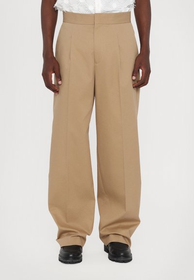 Beige weit geschnittene Hosen mit vorderen Falten, aus einem glatten Stoff gefertigt. Sie haben eine hohe Taille und einen entspannten Schnitt. Getragen mit schwarzen Schuhen.