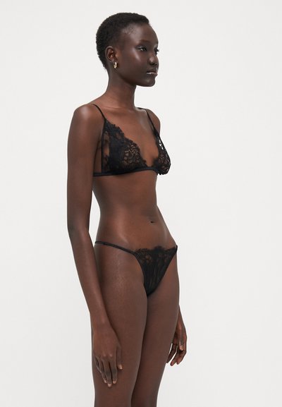 Conjunto de lencería de encaje negro que incluye un bralette y un tanga. Los delicados patrones florales, las tiras finas y la textura transparente realzan el diseño.
