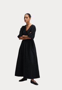 Schwarzes langes Kleid aus strukturiertem Stoff, mit V-Ausschnitt und Puffärmeln. Der Rock fällt knöchellang in lockerer Passform. Flache schwarze Schuhe.