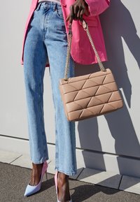 Person iført lyseblå jeans, lyserød blazer og pastelfarvede pink og blå højhælede sko, som holder en beige quiltet håndtaske med gyldne kædetræk.
