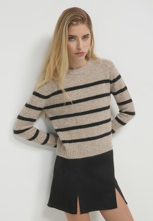 Donna con lunghi capelli biondi che indossa un maglione beige con righe orizzontali nere e una gonna nera con spacchi frontali, in posa contro un muro bianco.