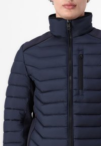 Jachetă puffer albastru marin cu guler înalt, fermoare și textură matlasată. Are un buzunar pe piept și accente negre pe umeri.