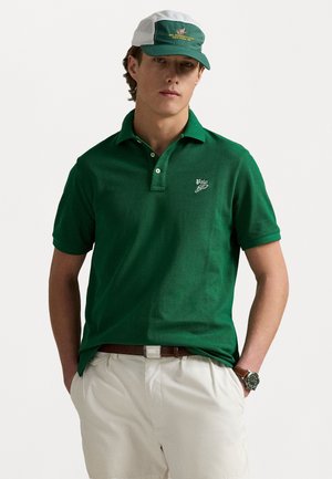 Grünes Poloshirt mit Kragen und kurzen Ärmeln, versehen mit einem kleinen Logo. Getragen mit hellen Shorts und einer grün-weißen Kappe.