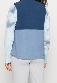 Osoba oblečená v dvoubarevném modrém fleecovém vestě přes dlouhý rukáv světle modrého trička s batikou a světle šedých kalhotách, zobrazená zezadu.