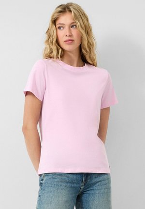 Frau mit welligem blondem Haar, die ein schlichtes, hellrosa T-Shirt mit kurzen Ärmeln und eine blaue Jeans trägt und leicht zur Seite schaut.