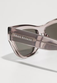 Occhiali da sole a forma di occhio di gatto con montatura traslucida rosa chiaro e lenti scure. Presenta il logo "PRIVÉ REVAUX" sull'asta in lettere argentate.
